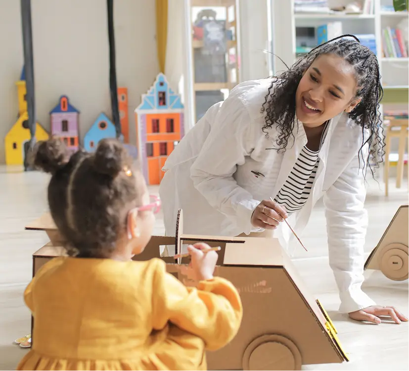 Smiling teacher leans over a classroom table to guide a child painting, creating a warm, encouraging art lesson with playful toys nearby